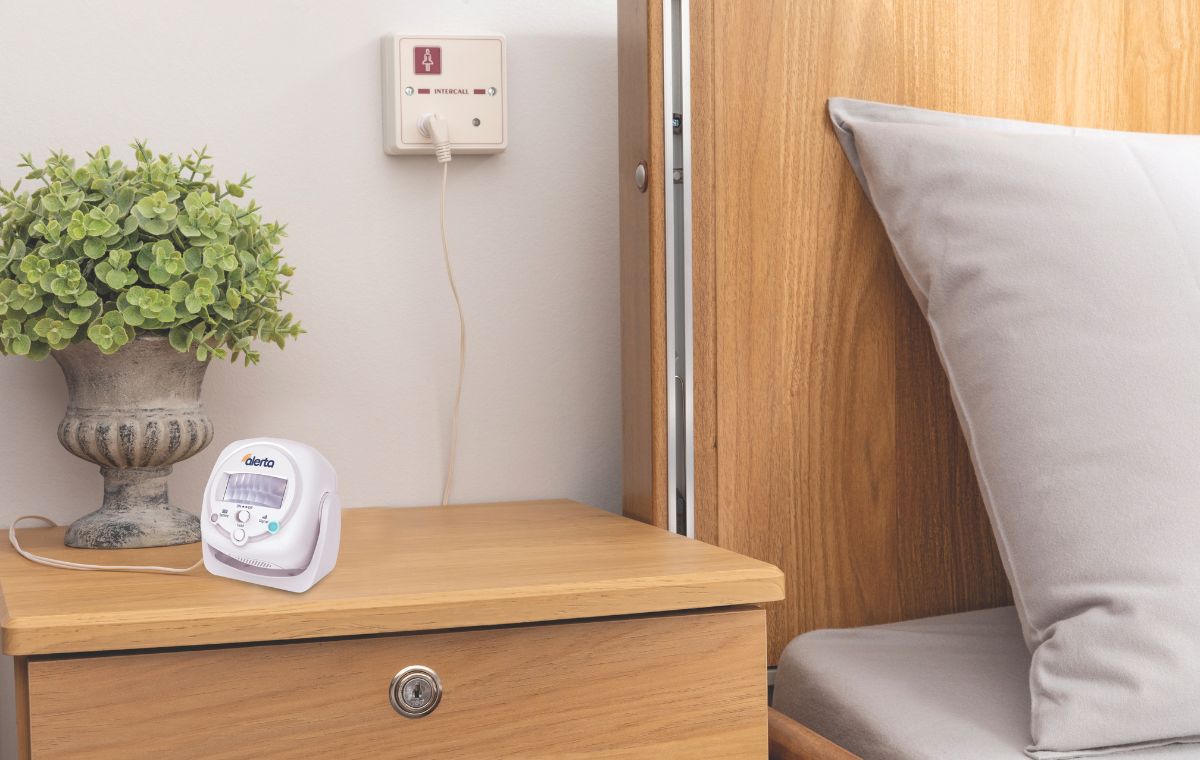 Alerta motion sensor on a wooden nightstand next to a bed with pillows and a plant.